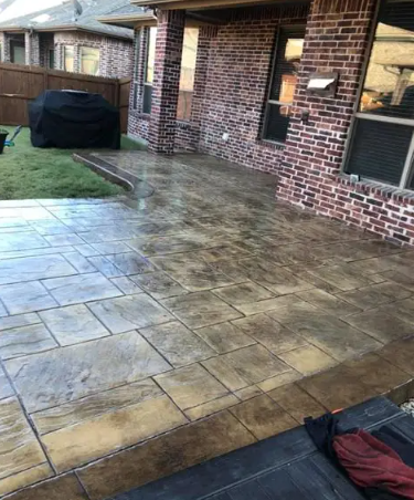 C&S Concrete Specialists - Backyard patio with stamped concrete slabs along a brick house; surface appears wet, with a covered grill and a grassy area nearby.